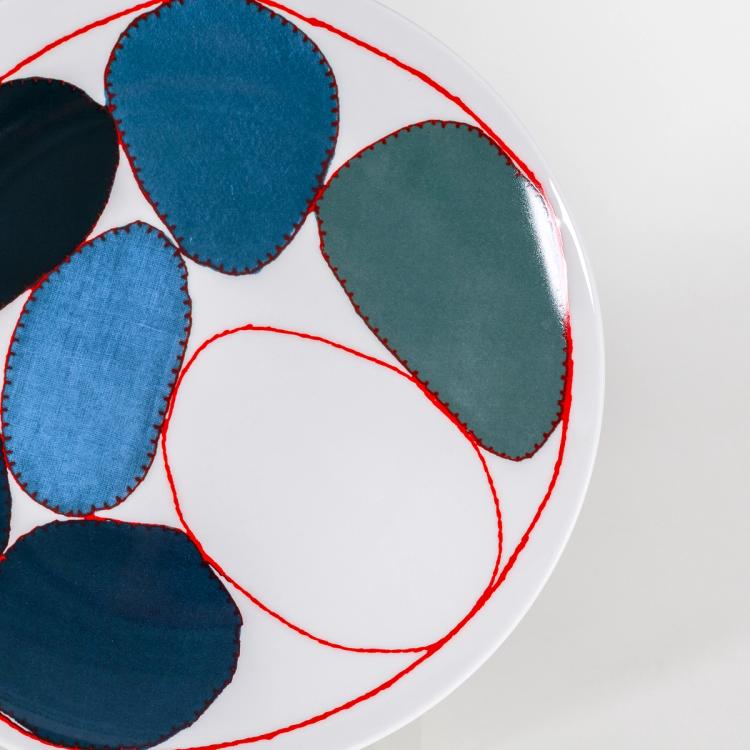 A detail photograph of the right edge to the center of a white plate with circles outlined in a red stitch design containing different fabric textures and shades of blue in the center. The circles are of varying sizes and shapes and they are encompassed in a larger red "stitched" circle bordering the outline of the plate.