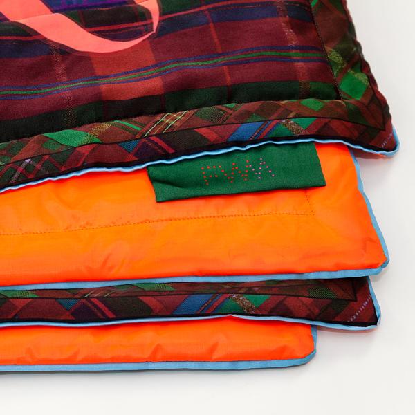 Four offset corners of a folded blanket show the top layer, with a plaid border of deep reds, greens, and blues, and its orange underside. A forest green label bears the FWM logo.