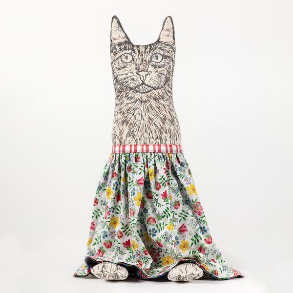 A photograph of a reversible flip doll. In this instance, a plush cat with drawn details stands upright with a skirt of florals screenprinted in warm, summery colors. The cat has little feet peeking out from beneath the skirt's hem.