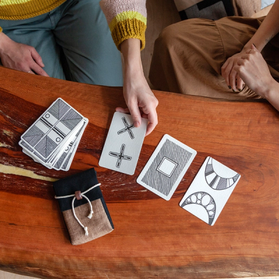 Two people are sitting at a wooden table. You can only see their hands and laps. The person sitting to the left is placing a card on the table, which has an "x" and a "+" design. It is sitting next to two other cards to the right that are laying down face up. To the left of the person's hand, there is a stack of cards sitting face down. In front of the cards, is a sewn pouch in black and brown linen with a white rope and dark brown clay bead closure.
