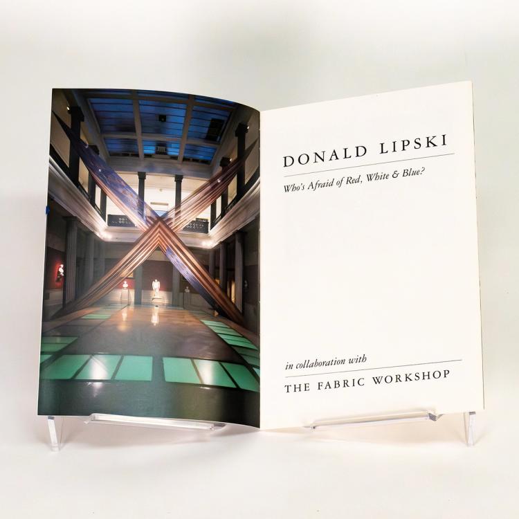 An open book spread featuring a color photograph in portrait orientation with an uneven black border. The photo is an exhibition view of Donald Lipski's sculpture titled Black by Popular Demand. The sculpture is of two 360 by 360 by 252-inch American flags assembled in an X formation, printed on silk organza fabric. The sculpture sits in an exhibition space at Corcoran Gallery of Art. The space has two stories. Grey floors with blur squares assembled in a rectangle, and is dimly lit.