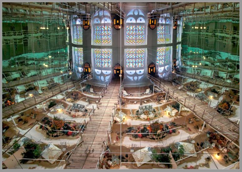 A full-bleed book spread featuring an art installation consisting of a refracted architectural space ending with a repeated and mirrored stained glass window. The lower refractions are divided by wooden boardwalks with smaller clusters of sand, furniture, and assorted objects.
