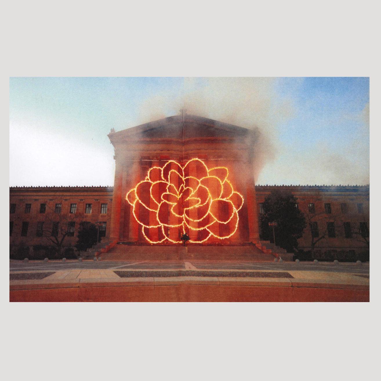 An image of a book spread featuring one full bleed photograph. The photo is of the Philadelphia Museum of Art, a grand building with columns on a blue-sky day. In front of the columns is an explosion in the shape of a flower (like a firework) with lots of hazy smoke around it. 