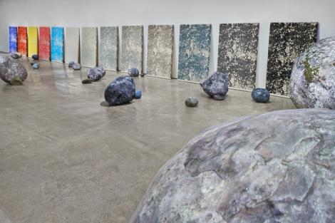 An installation view of a contemporary art exhibition featuring 13 screenprinted panels leaned against a wall. Each panel features the same image of a rough texture but is printed in a different colorway, stretching from primary colors to warm and cool grays. Sculptural boulders and smaller rocks are placed along the gallery floor.