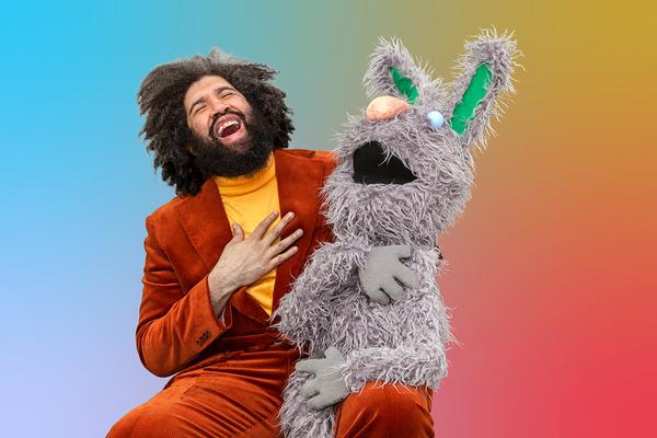 A man and a large rabbit puppet give a hearty laugh with their hands on their chests. The man is wearing a burgundy corduroy suit with a yellow turtleneck. He has thick dark curly hair that blends in with his beard. The rabbit puppet is grey with stringy fur, a round sky blue eyeball, a pink nose, and green at the center of its tall ears.