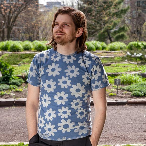 A model stands outdoors wearing a sky blue t-shirt with a series of white daisy-like flowers printed across it.