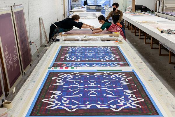 A photograph of a long table receding in perspective at the top of the image. Two sets of long rectangular patterned designs appear in the foreground. Further back, three artists work from both sides of the table. Two of them are leaning over to screenprint together.