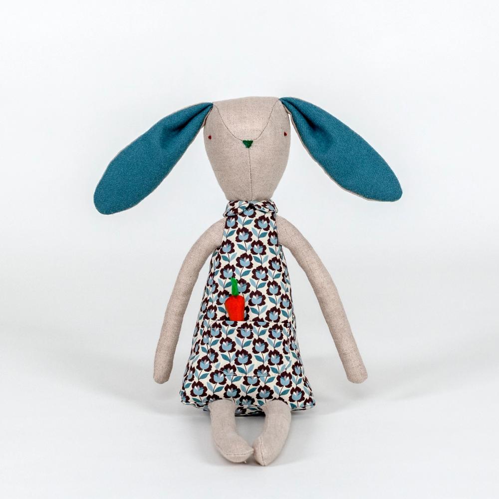 A photograph of a plush rabbit made of light beige woven fabric characterized to have a pointy nose, triangular shaped body, and long thin arms and legs. The body of the rabbit is made of a primarily blue patterned fabric and includes a pocket with a removable plush orange carrot.