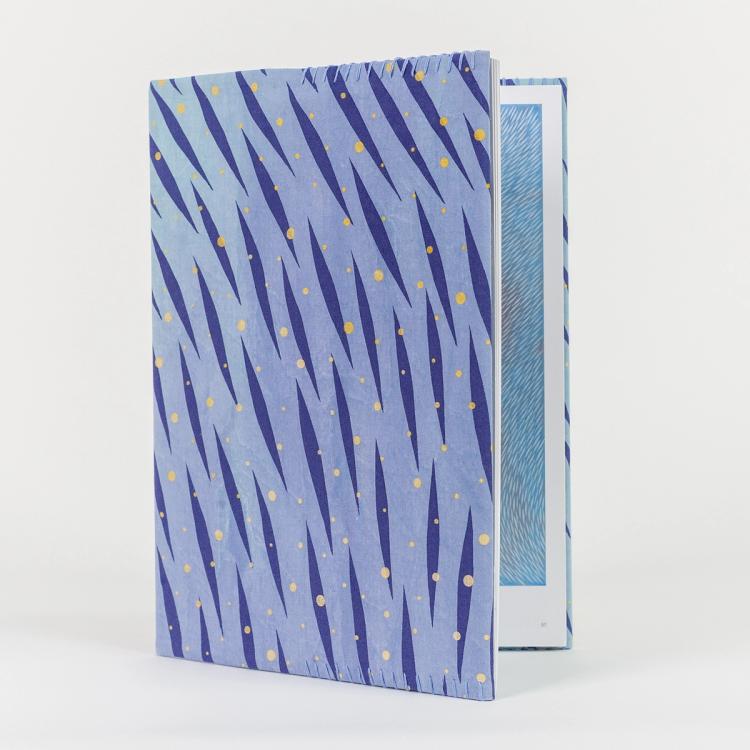 A photo of a hardback book with a hand-stitched book cover featuring a vibrant pattern of diagonal dark blue brush-like streaks and scattered yellow dots on a light blue background. The book is slightly open, revealing a blue patterned illustration inside.