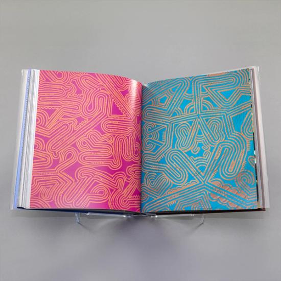 A photo of a book spread with a full-bleed photograph of two labyrinthian designs by the Maisin Artists of Papua New Guinea on each page. The one on the left features a yellow design over a hot pink ground; on the right is a yellow design over a cerulean blue ground.