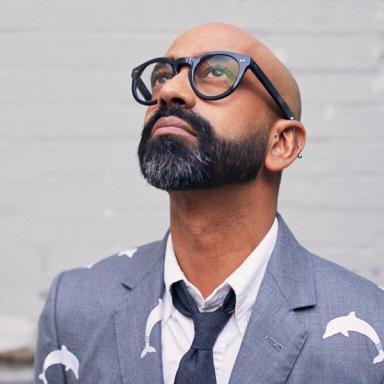 A portrait of the artist Brendan Fernandes, a brown-skinned man with a shaved head and black beard. He is looking up while wearing black-rimmed glasses, a blue-gray suit, and gray tie.