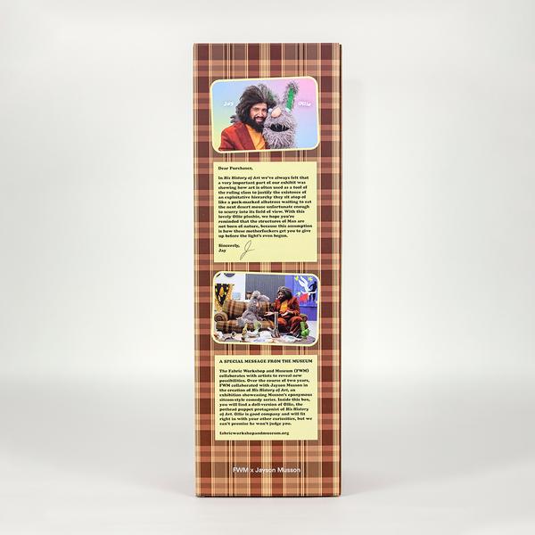 A photo of a tall brown plaid box featuring two images of a man with dark bushy hair wearing a burgundy coat and yellow turtleneck with a scraggily gray rabbit. The image on top features the two posing together while the image below features the two on a brown plaid couch similar to the box. Each image is accompanied with a yellow message. The lower register of the box reads "FWM x Jayson Musson"