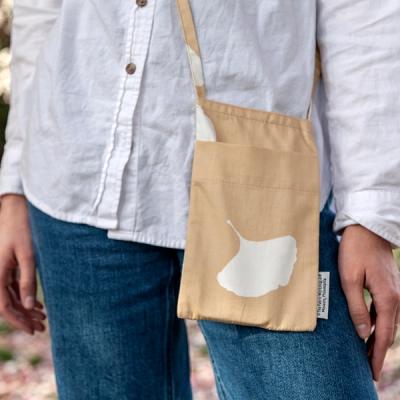 A close up photo of a model's hip wearing a small, rectangular, cross-body bag. The bag is tan with a life-size, white silhouette of a ginkgo leaf. There is a small label tag on the right side seam of the bag.