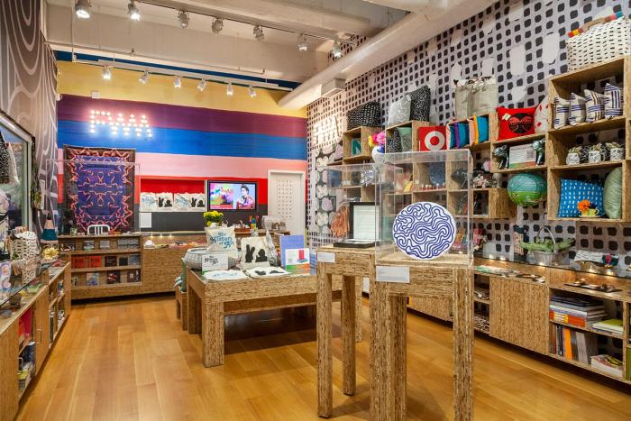 A photograph of The Fabric Workshop and Museum store. The walls are covered floor to ceiling in patterned wallpaper. On the far wall are stripes of purple, blue, pink, red, and black. "FWM" is spelled out in lights. On the wall to the right are a number of wooden shelves and cabinets with various colorful products displayed. Several sets of display fixtures both tall and skinny as well as broad and low feature additional products, some under plexiglass protection like a plate by the artist Sol Lewitt that features a blue squiggly drawing. 