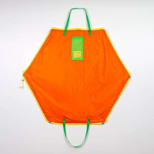 A hexagonally-shaped orange bag hangs on a wall. It features two green straps, a green pocket, and yellow tag that reads "Risa Puno x FWM." The bag is outlined in yellow nylon.