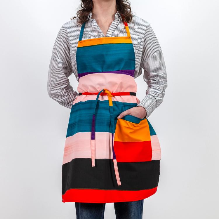 A model wearing a long tie-apron featuring painted horizontal stripes of purple, light pink, teal, red, black, and ochre. The model has her hand placed in the front pocket of the apron