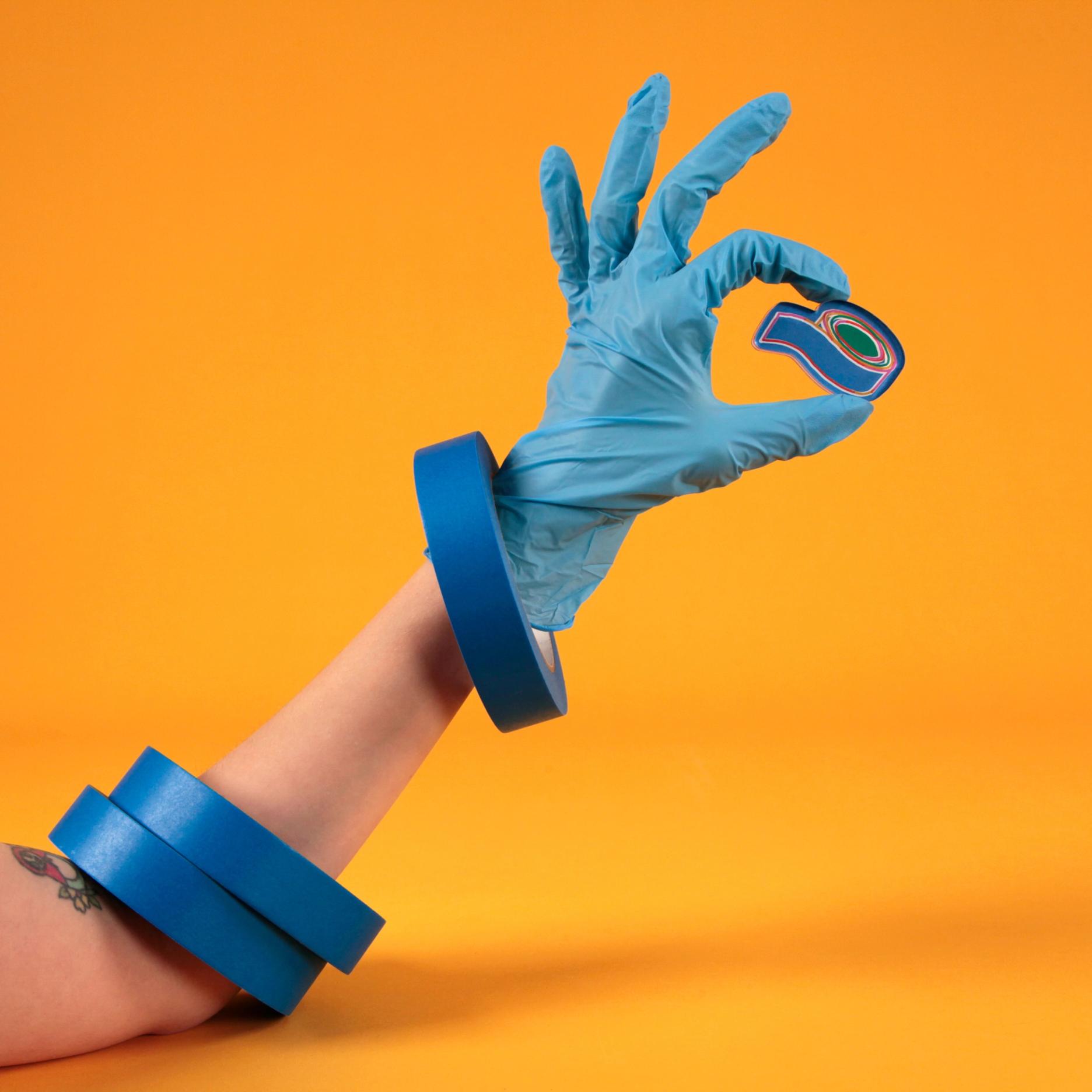 A hand wearing a blue glove with three rolls of blue tape around the wrist like bracelets is holding the blue tape magnet against a bright, warm yellow background.