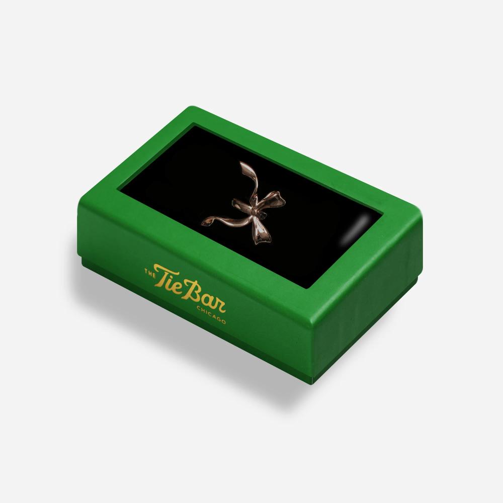 A rectangular box with a green border features a rose gold colored scarf clip shaped like a bow inside. The side of the box reads "The Tie Bar, Chicago"