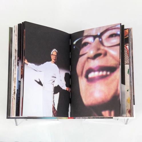 A book spread showing on the left page an older white woman in a minimally designed long-sleeve white dress smiling at the viewer. She appears lit as if on a stage with her shadow behind her. On the right is an extreme close-up with a soft focus of her smiling face at a three-quarter angle, 