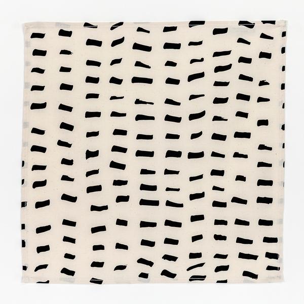 A square napkin composed of cream-colored fabric with several rows of short brushstroke-like black dashes.