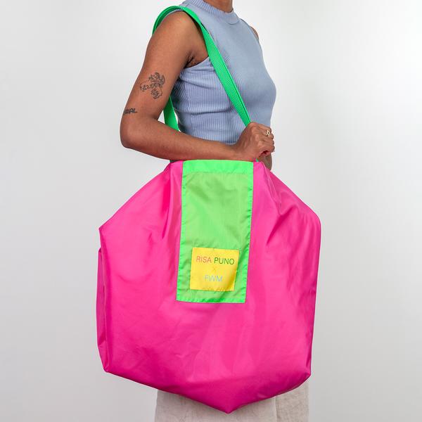 A person holds a large nylon bag on their shoulder. The bag is magenta with green straps and a yellow tag that reads "Risa Puno x FWM."