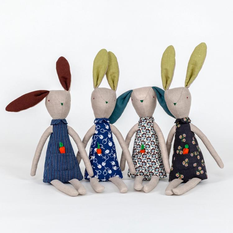 A photograph of a grouping of four plush rabbits made of light beige woven fabric characterized to have a pointy nose, triangular shaped body, and long thin arms and legs. Each have a body made of a different blue patterned fabric and includes a pocket with a removable plush orange carrot.