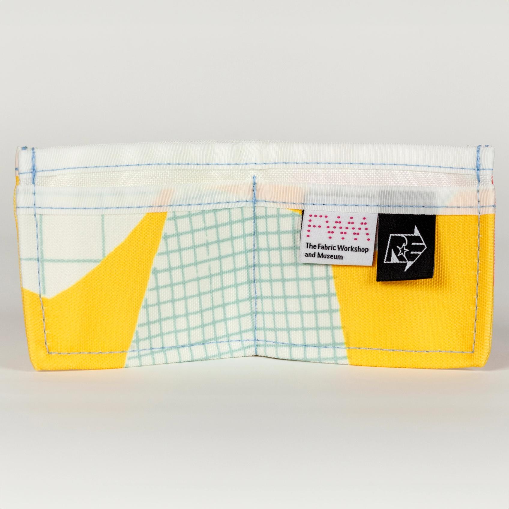 An open vibrant yellow wallet (opens into a wide rectangle with one large pocket in the back and two smaller pockets in front) with bold abstract shapes (white with a light blue grid pattern). There is a solid white seam lining the top of the wallet. There are two tags on the right pocket. The tag on the left is white with a pink dotted logo reading "FWM" with "The Fabric Workshop and Museum" written underneath. The tag on the right is black with an arrow. Inside the arrow are the letters "RE"