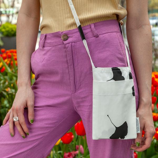 A close up photo of a model's hip wearing a small, rectangular, cross-body bag. The bag is white with a life-size black silhouette of a ginkgo leaf. There is a small label tag on the right side seam of the bag.