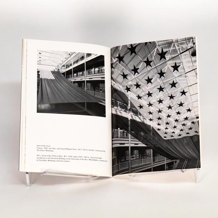 A photograph of an open spread of a book. There are two black and white photos of different angles of the same art installation. The photos are an exhibition view of Donald Lipski's sculpture titled Who's Afraid of Red, White & Blue #31. The sculpture is an 840 by 288-inch American flag printed on translucent nylon. The flag has been installed in a glass ceiling atrium at the Haviland Building of Philadelphia's University of the Arts.