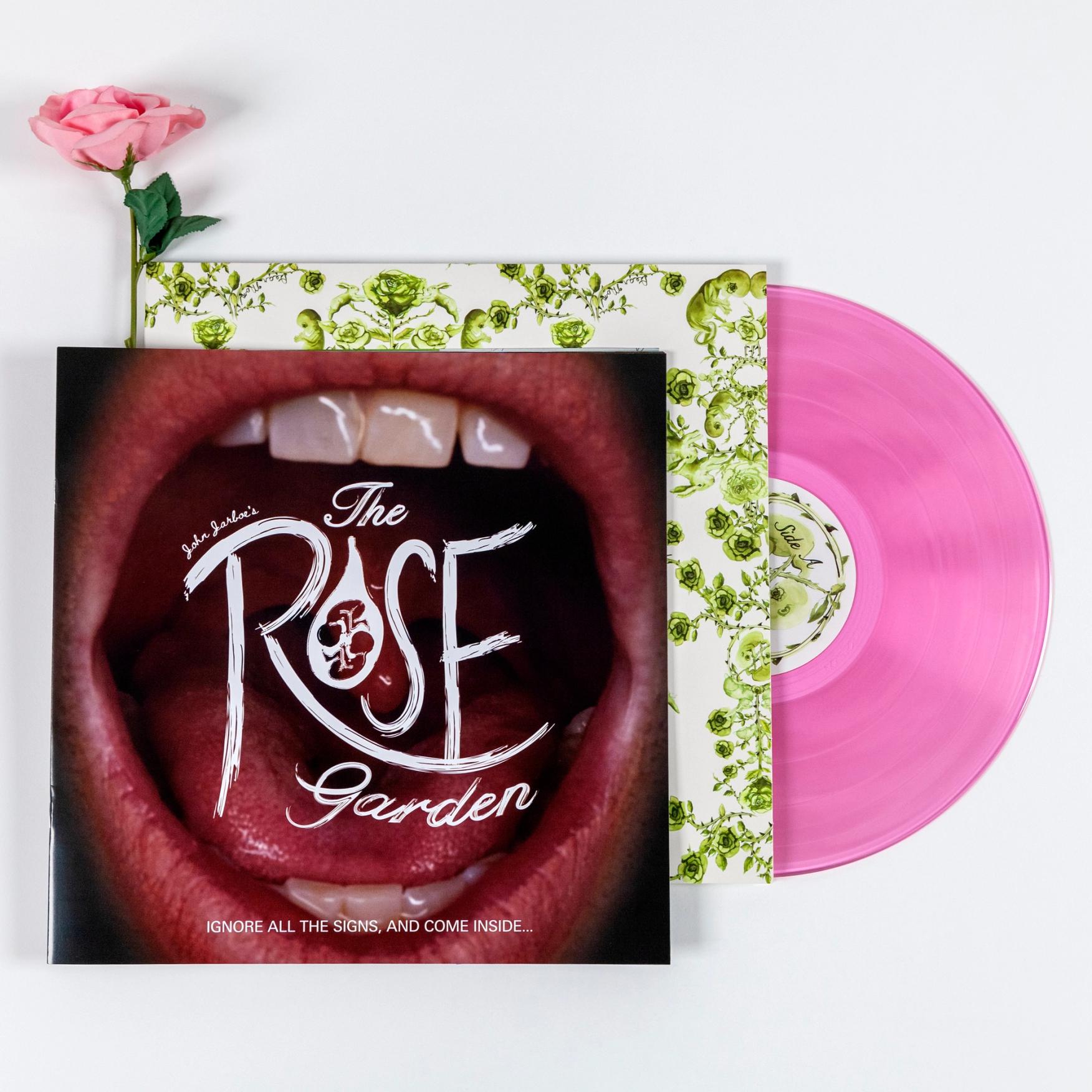 A cover of a square book laying on top of a record cover with a pink see-through vinyl coming out of the cover. The image on the book is a close-up photo of a mouth open wide. There is text overlaying the open mouth that reads "John Jarboe's The Rose Garden IGNORE ALL THE SIGNS, AND COME INSIDE..." There is a pink rose poking out from behind the top of the book.