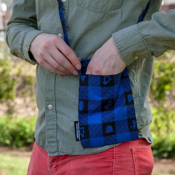 A model reaches into a small flat bag at his hip. The bag features a blue and black plaid pattern with icons of objects in the black squares.