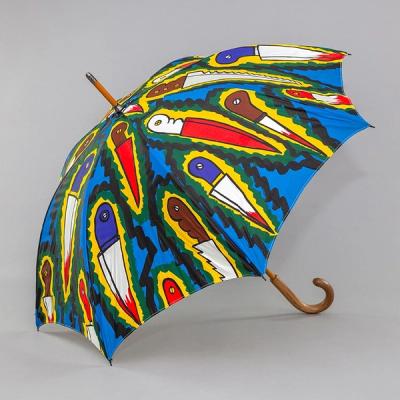 An umbrella sits against a grey background. It is bright and colorful, with pop art style knives with bright red blades and tips, outlined in electric yellow with a dark zigzag and sitting against a bright blue background. The umbrella has a curved wooden handle.
