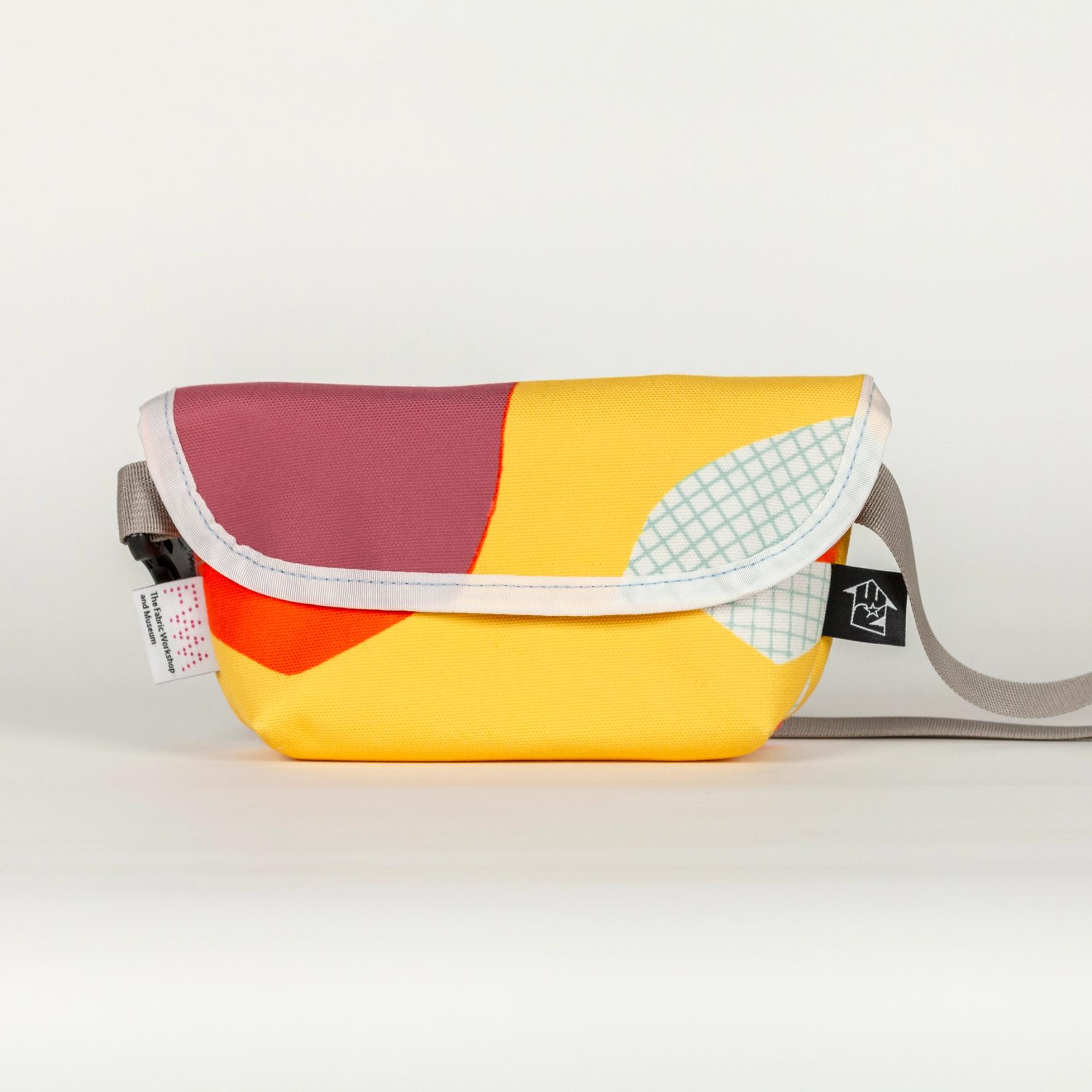 A short, wide, vibrant yellow bag with bold abstract shapes (bright orange, purple, and white with a light blue grid pattern). The bag has a velcro top flap closure with a white seam along the flap. The bag has a shiny gray strap and two tags; one on the left and one on the right. The tag on the left is white with a pink dotted logo reading "FWM" with "The Fabric Workshop and Museum" written underneath. The tag on the right is black with an arrow. Inside the arrow are the letters "RE"