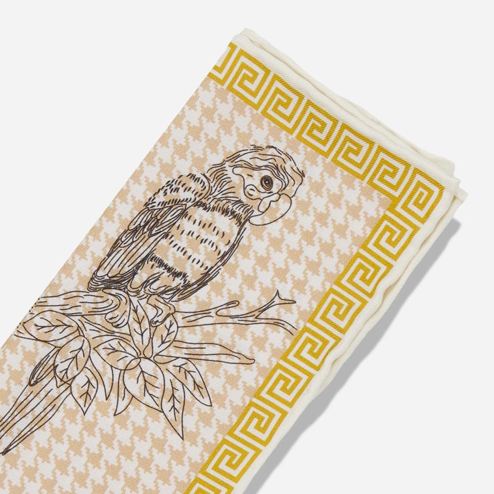 A folded pocket square with a detail of the corner. Its gold border resembles a Greek-style spiral frieze. There is a black line illustration of a Kasuku parrot sitting on a leafy branch on a light pink and white houndstooth background.