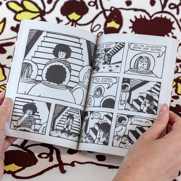 A photograph of two hands spreading a book's pages against fabric yardage consisting of brown and yellow floral motifs. The book features cartoon panels with women astronauts arguing over what to do about a distraught woman they've encountered who is not wearing a spacesuit.