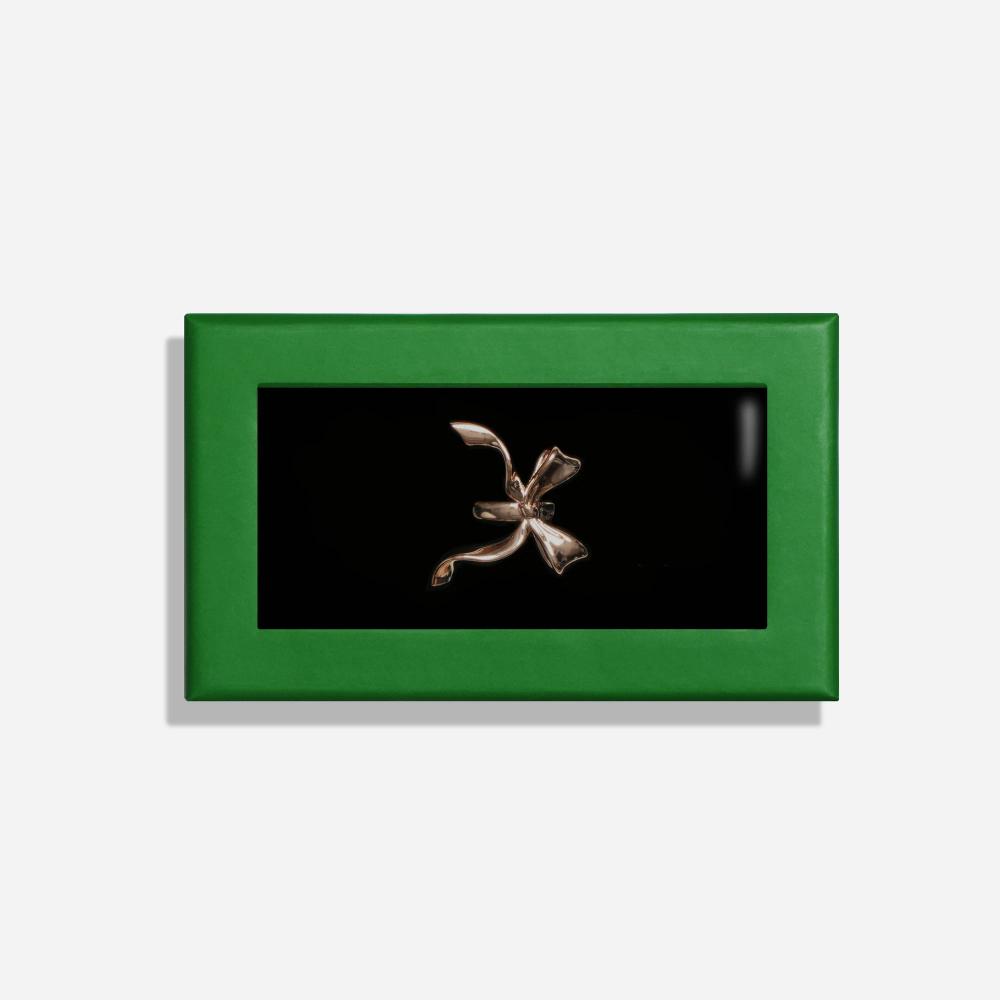 A rectangular box with a green border features a rose gold colored scarf clip shaped like a bow inside.