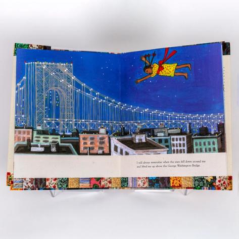 A photograph of an open spread of a picture book by artist Faith Ringgold. The same illustration takes up both pages. The image depicts a whimsical scene of a night sky, tall buildings taking up the skyline, and the George Washington Bridge. There is a girl in a yellow dress flying above the buildings.