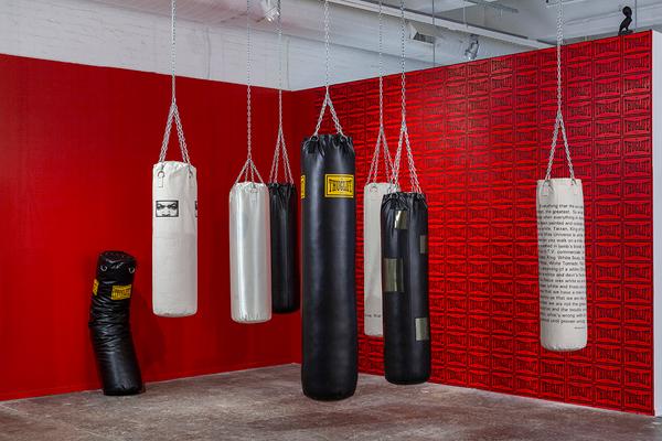 A photograph of a gallery installation featuring a set of seven punching bags suspended against two red walls with one additional punching bag slumped against the left wall. The bags vary in height and color (four are white, four are black). The central bag, which is the tallest, features a yellow graphic that reads "Thuglife" stamped over black fabric. The same graphic repeats as red wallpaper behind it.