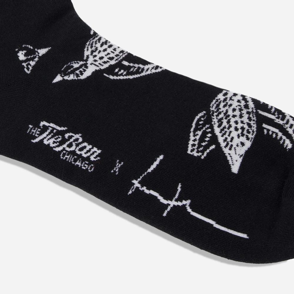 A black knit dress sock with a pattern of white illustrated Shomoro birds flying in different directions. The artist's signature and "The Tie Bar" logo is featured on the bottom of the sock.