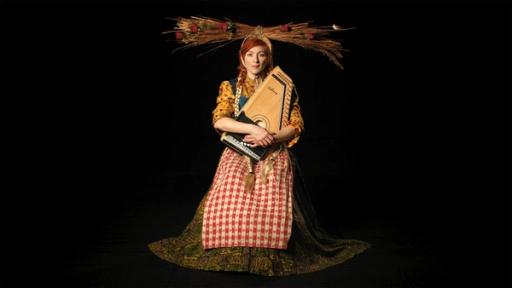 A photograph of a kneeling woman theatrically lit in a pitch black environment. She is wearing straw carefully balanced on her head, a yellow floral shirt with rolled up sleeves, a long brown dress, and a red plaid apron. She is holding with both arms a zither, a kind of string instrument.
