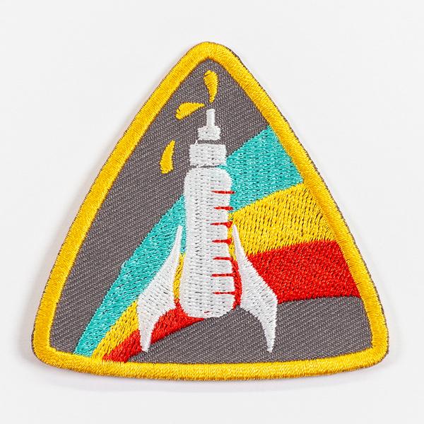 A triangular patch with rounded edges outlined in a thick yellow border. At its center is an embroidered white bottle with wings so as to resemble a rocket ship. The bottle's tip is emitting three drops of liquid. Three colored bands streak behind the bottle rocket. 