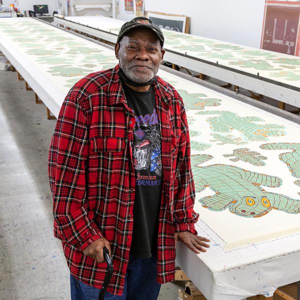 A picture of Will Stokes, Jr., an older Black man, smiling for the camera. He is resting his weight on a cane with his right hand and on a long print table with his left hand. He is wearing a black cap and a large red plaid shirt with a black t-shirt underneath. On two long printmaking tables are a series of repeated shapes representing an alligator