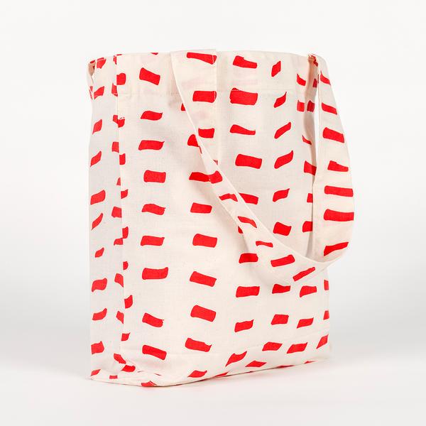 A tall, rectangular tote bag made of white fabric yardage with a printed pattern of short, gestural, brushstroke-like red dashes.