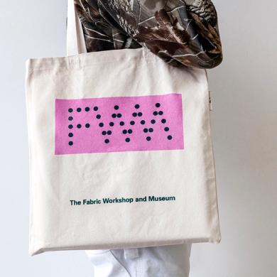 A photo of a model wearing a light tan canvas tote bag. On the bag, there is a screenprinted pink rectangle (landscape). Inside the pink rectangle are the letters "FWM" and the letters are made up of bold, dark green dots. Underneath the rectangle in a smaller, bold, dark green font reads "The Fabric Workshop and Museum"