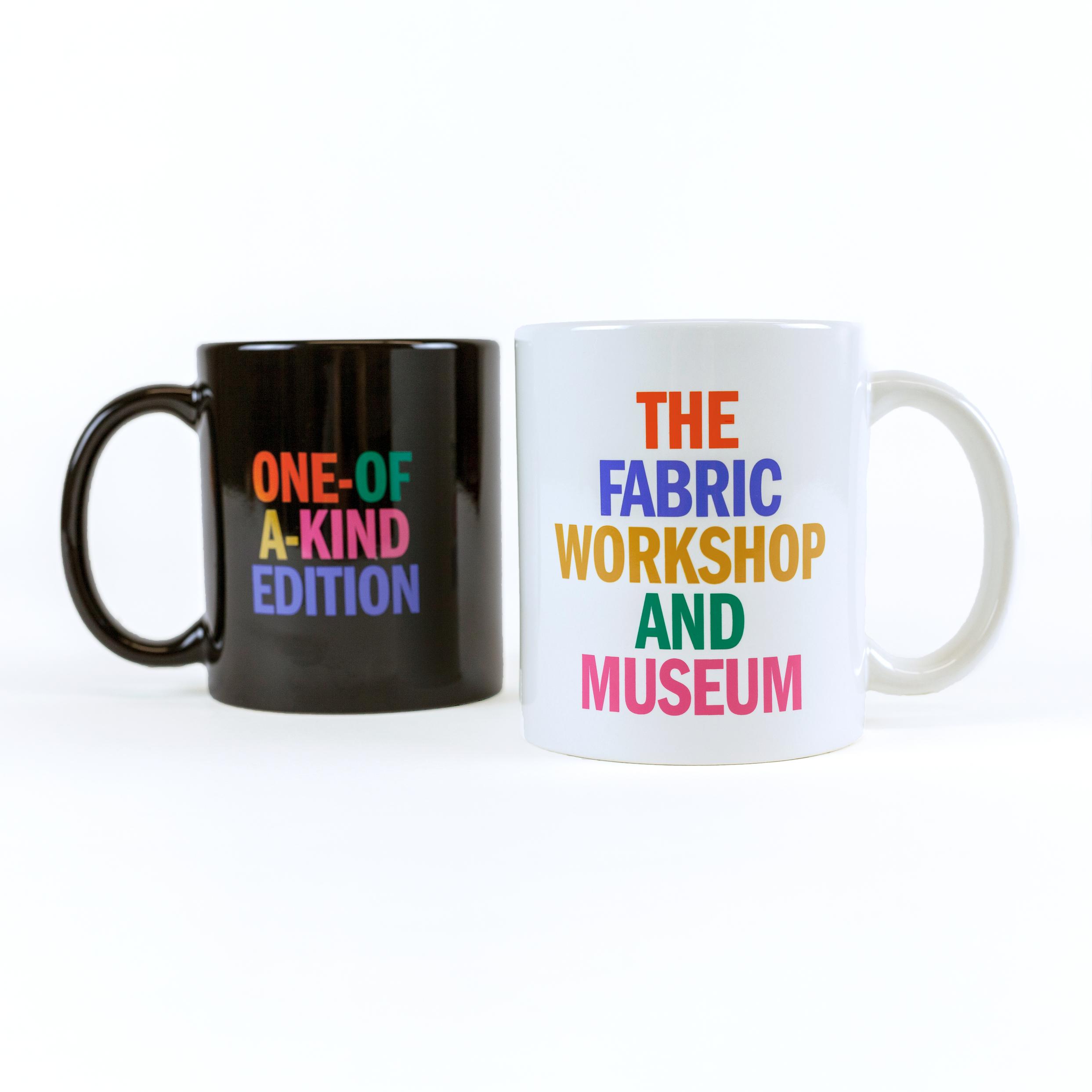 Two ceramic mugs, one black and the other cream colored, sit next to one another against a white background. The black mug, slightly further back, reads "ONE-OF-A-KIND EDITION". Each word is in a different color: orange, green, gold, pink and blue. The white mug, with its handle facing opposite, reads "THE FABRIC WORKSHOP AND MUSEUM." Each word is stacked and in a different color: orange, blue, gold, green, and pink.