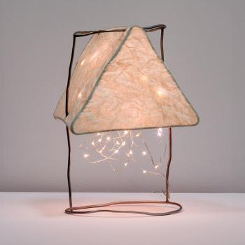 An upright paper lamp glows warmly from within a triangular armature with string lights spilling out below. It is suspended above a table surface from two crooked rods that bridge two circular-shaped rods.