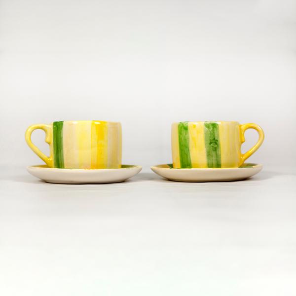 Two cups and saucers sit side by side against a white backdrop—they each have slightly varying wide vertical stripe patterns in shades of yellow and green. 