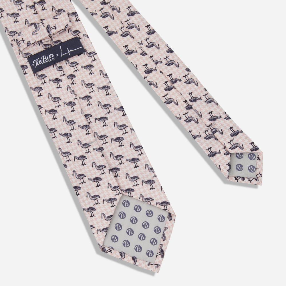 A detail of the back of a tie with a pattern of black illustrated Bata birds with long beaks in alternating stances on a light pink and white houndstooth background. There is a label with the artist's signature and "The Tie Bar" logo.