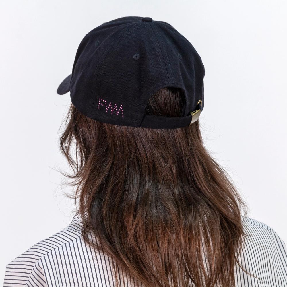 A photo of the backside of a model wearing a black baseball cap. There is a pink dotted logo spelling out "FWM" on the left side of the adjustable strap.