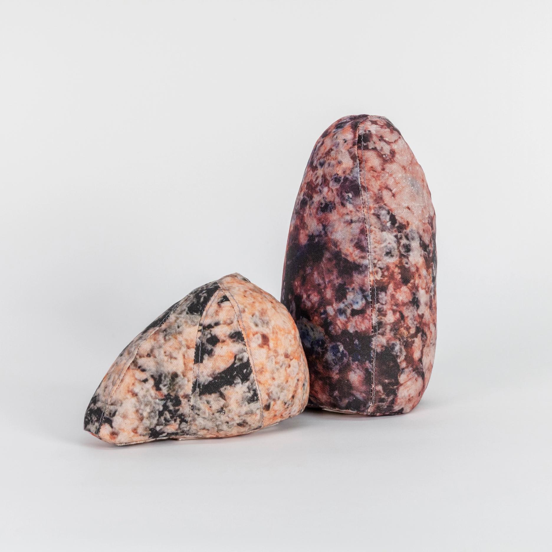 A photograph of two small soft sculptures covered with patterns that resemble polished stone of different hues. One sculpture is short and triangular where the other one is tall like an obelisk with a rounded top.